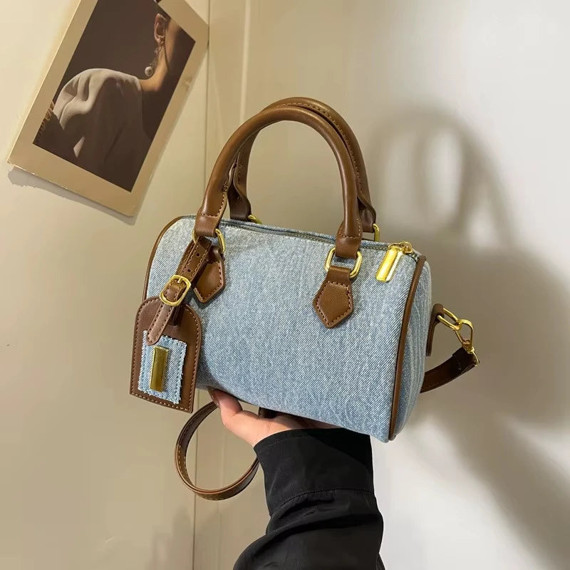 Light blue denim cylinder handbag with brown leather trim, gold hardware, and matching luggage tag. Designer style.
