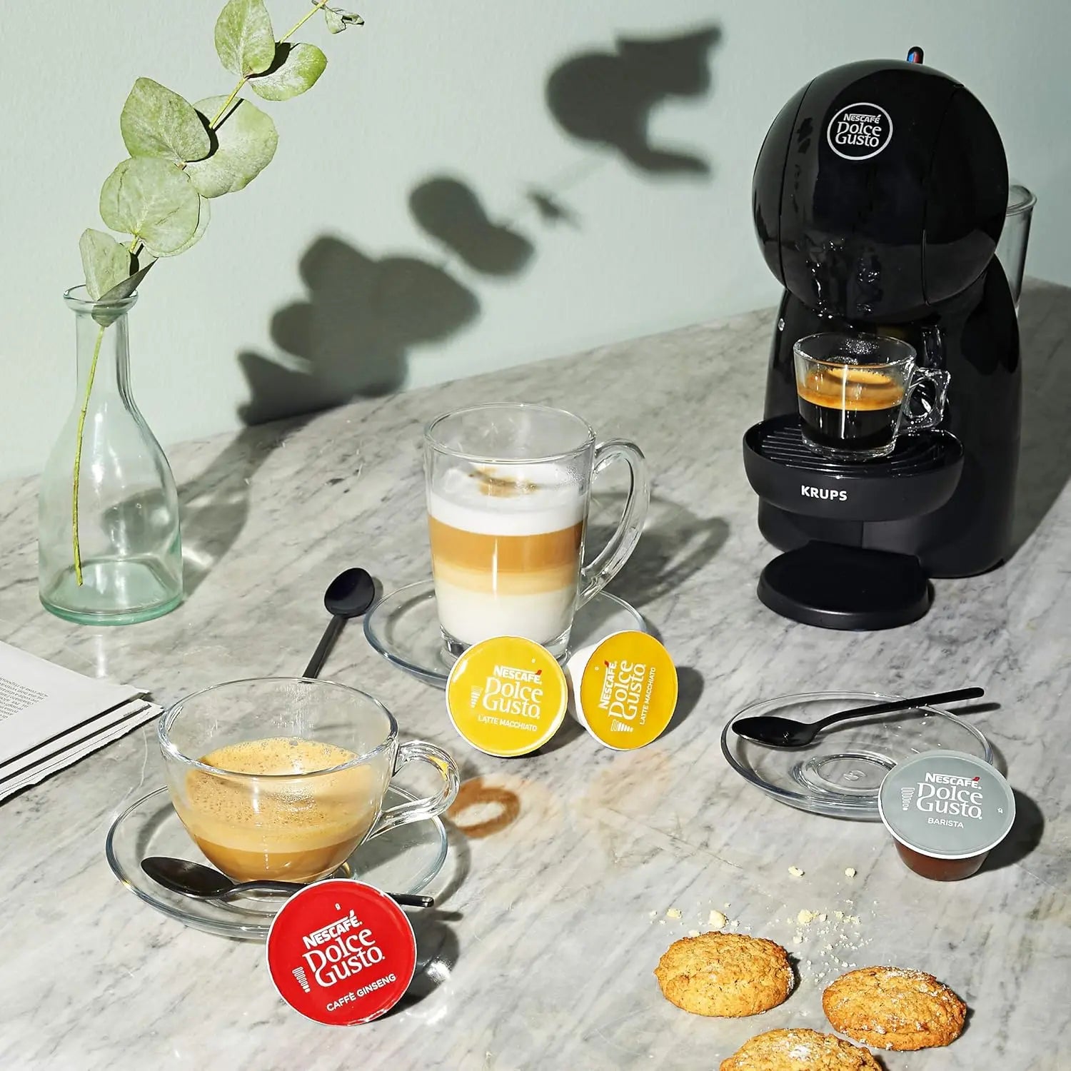Black Krups Nescafé Dolce Gusto capsule coffee machine with glass mugs and assorted coffee pods on marble surface.
