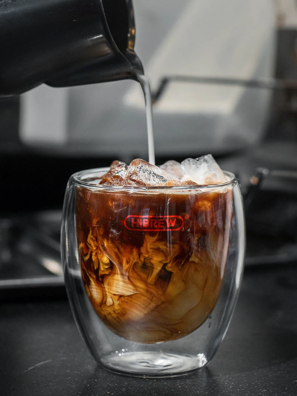 HiBREW branded clear double-walled glass cup being filled with iced coffee and milk.