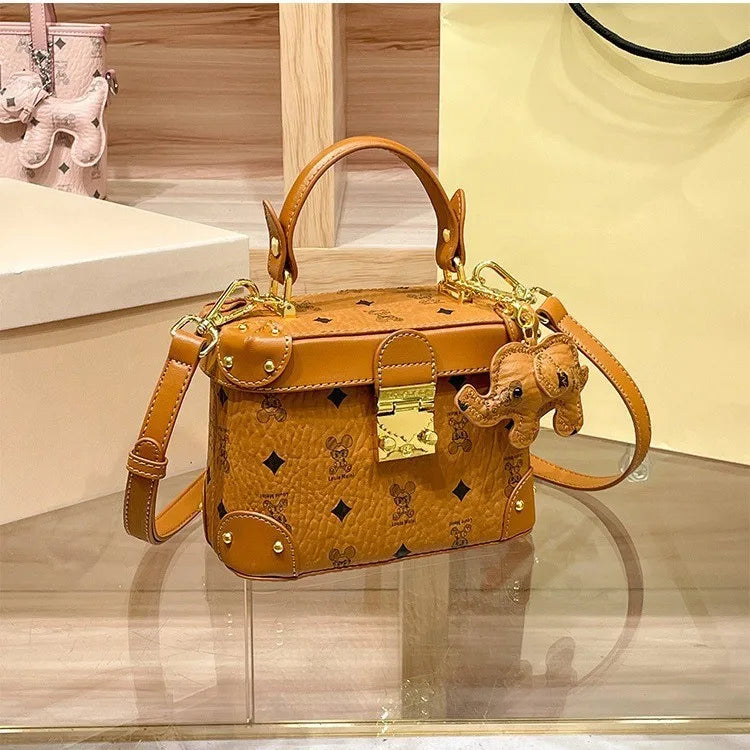 Tan cowhide trunk handbag with gold hardware, monogram print, and elephant charm. Minimalist chic style for women.