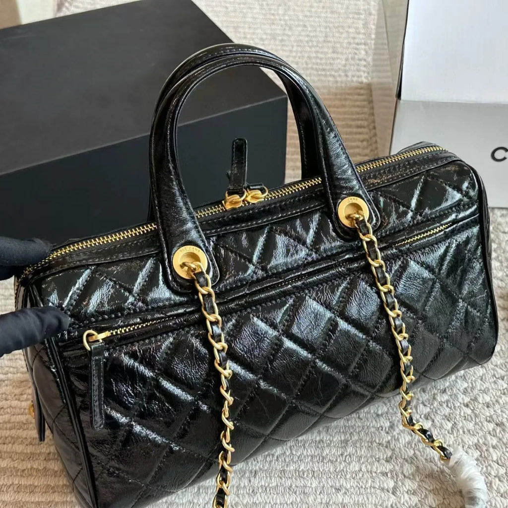 Black patent leather diamond-quilted barrel satchel with gold chain-link straps and zip hardware. Designer handbag.