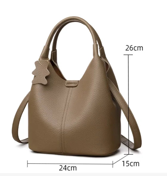 Taupe pebbled leather bucket bag with top handles, crossbody strap, and cute bear charm for casual daily commuting.