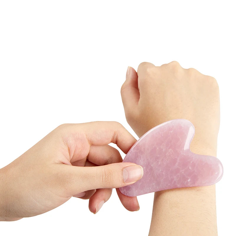 Hand holding a smooth, heart-shaped rose quartz gua sha tool against a wrist, isolated on a white background.