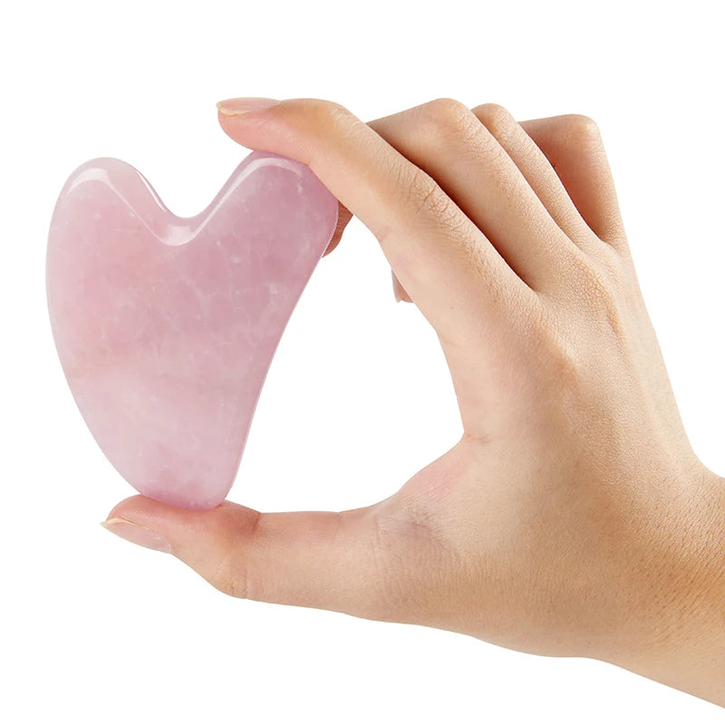 A hand holding a heart-shaped, smooth, pale pink rose quartz gua sha tool for facial massage.