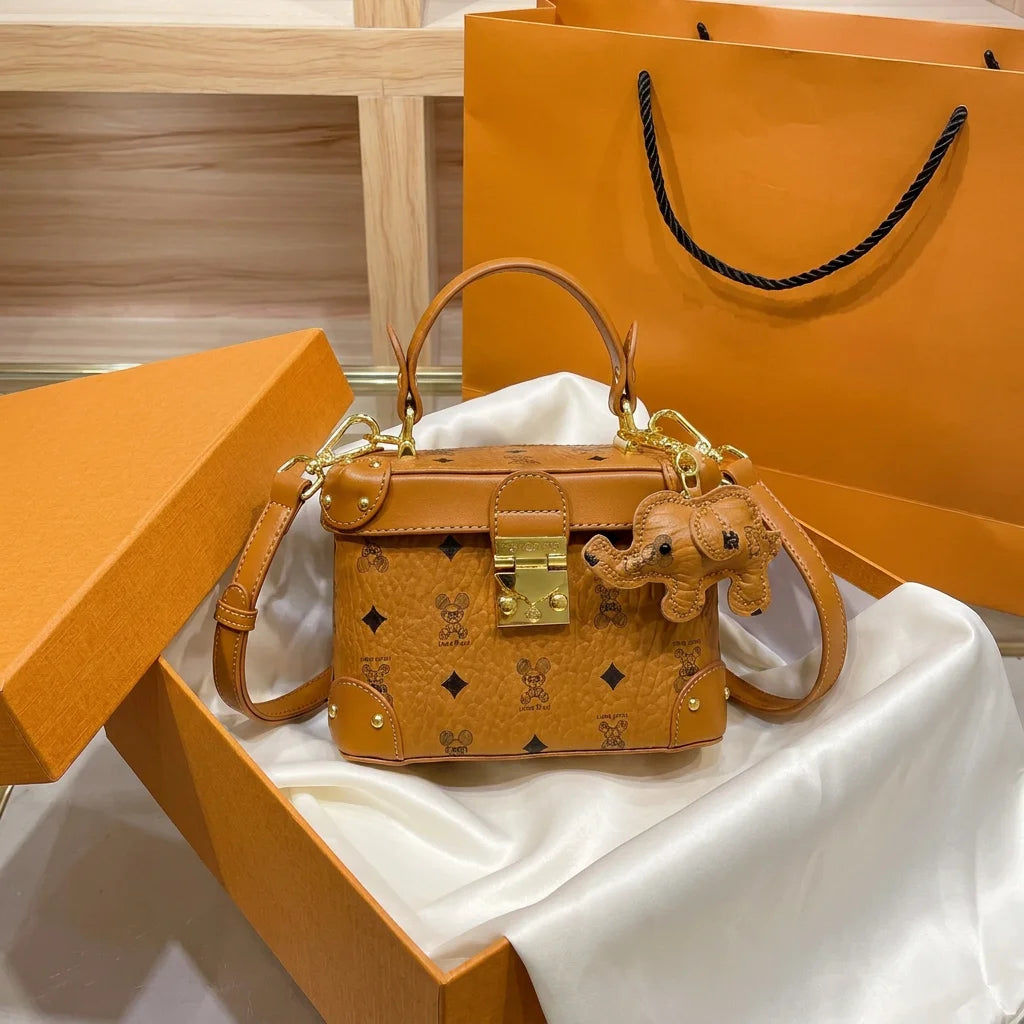 Tan cowhide leather trunk handbag with monogram print, gold hardware, top handle, and cute elephant bag charm.