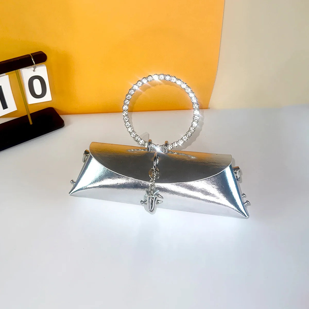 Silver metallic PU clutch with crystal-encrusted round metal handle, ideal for luxury evening or wedding wear.