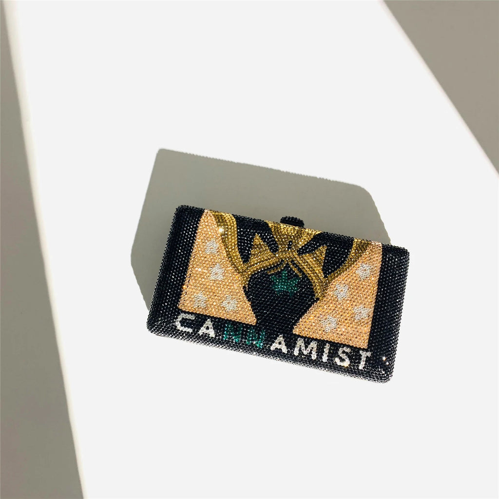 Crystal-embellished black designer clutch bag with gold "CANNAMIST" logo and decorative crown motif for evening wear.
