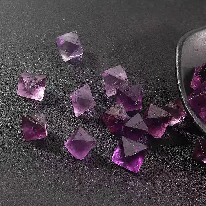 Small purple rough crystal octahedra on dark surface, some near metal spoon