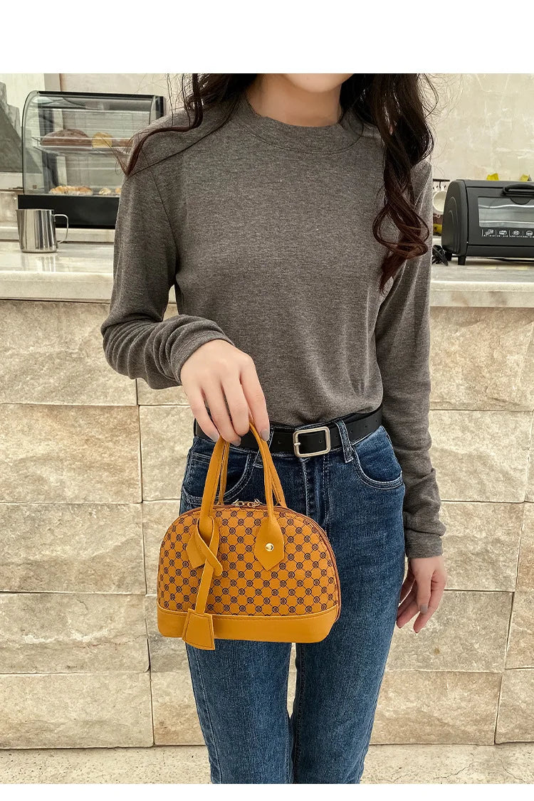 Mustard yellow designer shell handbag with dark brown monogram print, dual top handles, and secure zip closure.