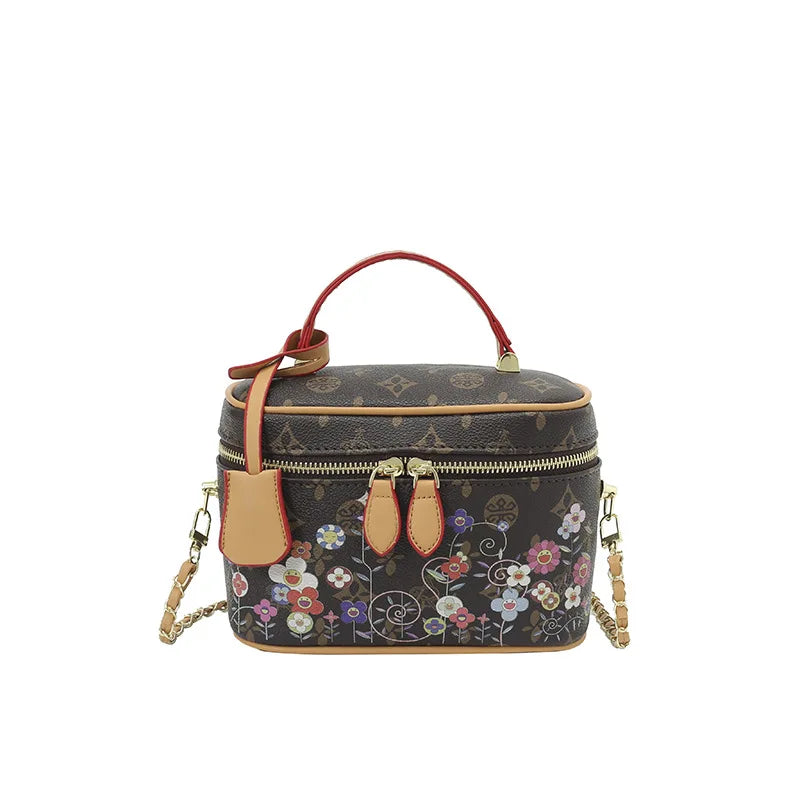 Brown leather floral trunk crossbody bag with red handle, gold chain strap, and zipper closure.