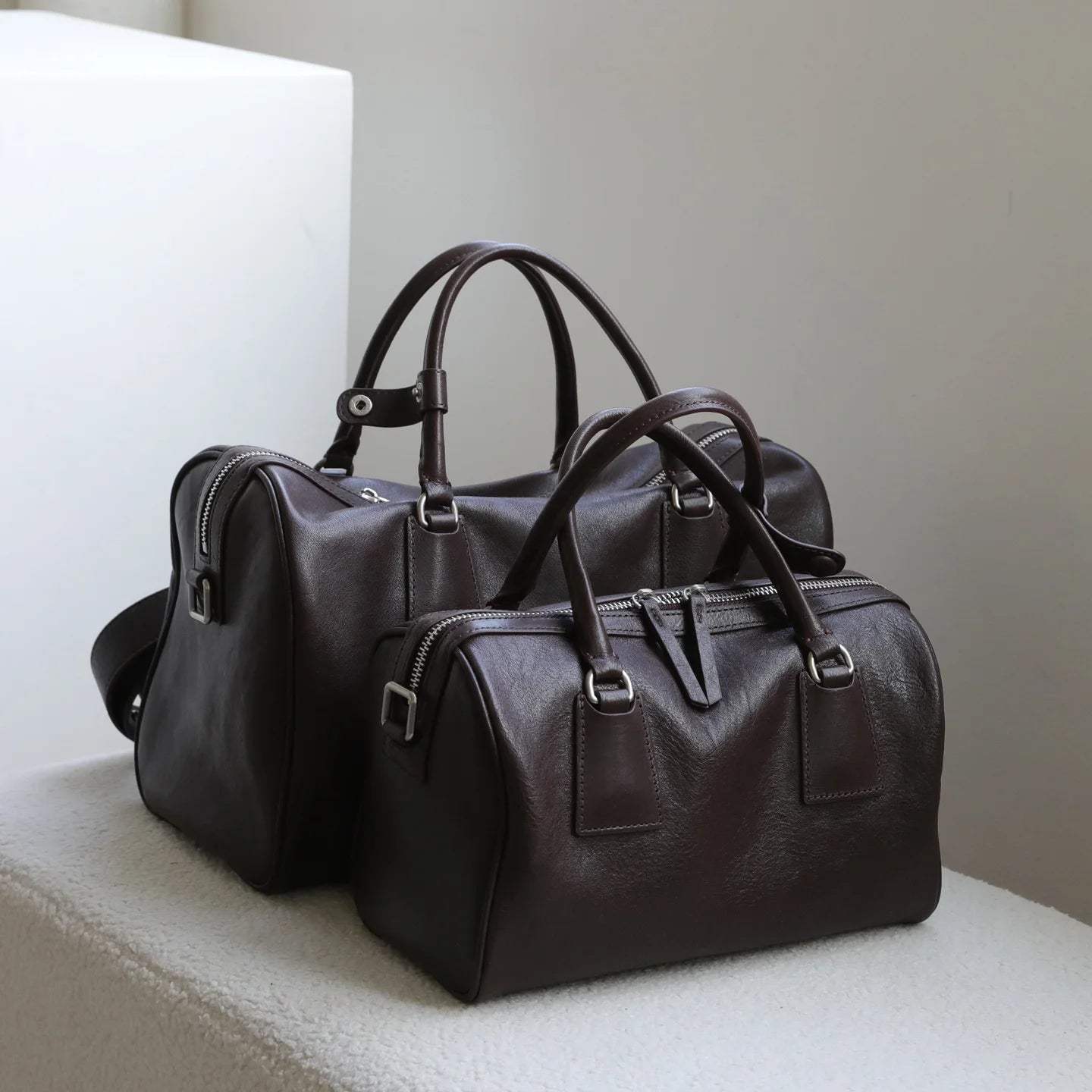 Two dark brown vegetable-tanned leather Boston bags with double zip closures and minimalist designer aesthetic.