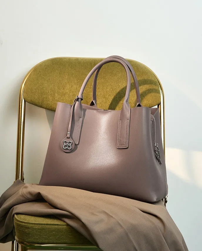 Mauve genuine leather designer handbag with dual handles, side zipper, and clover charm on a green velvet chair.