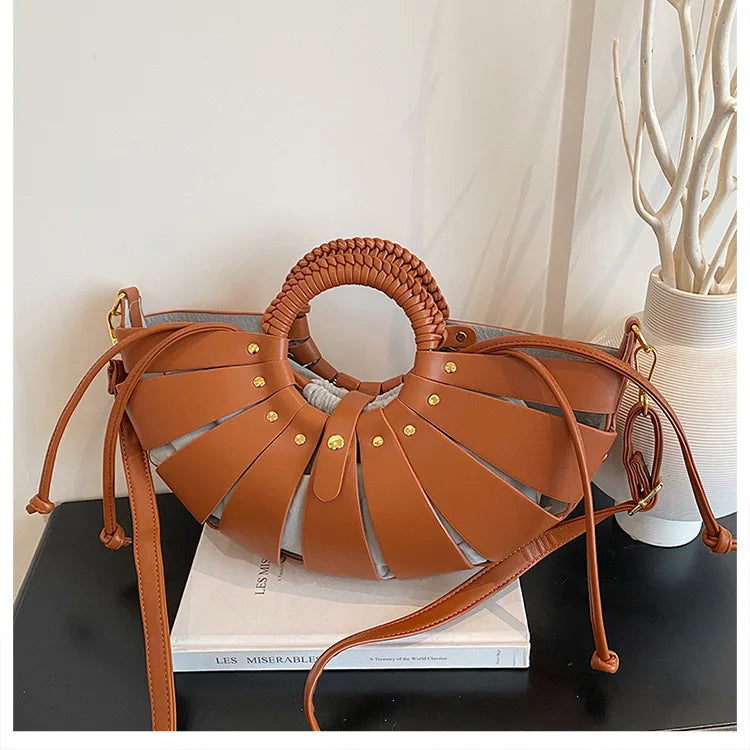 Tan half-moon crossbody trunk bag with hollow-out leather strips, braided round handle, and gold stud details.