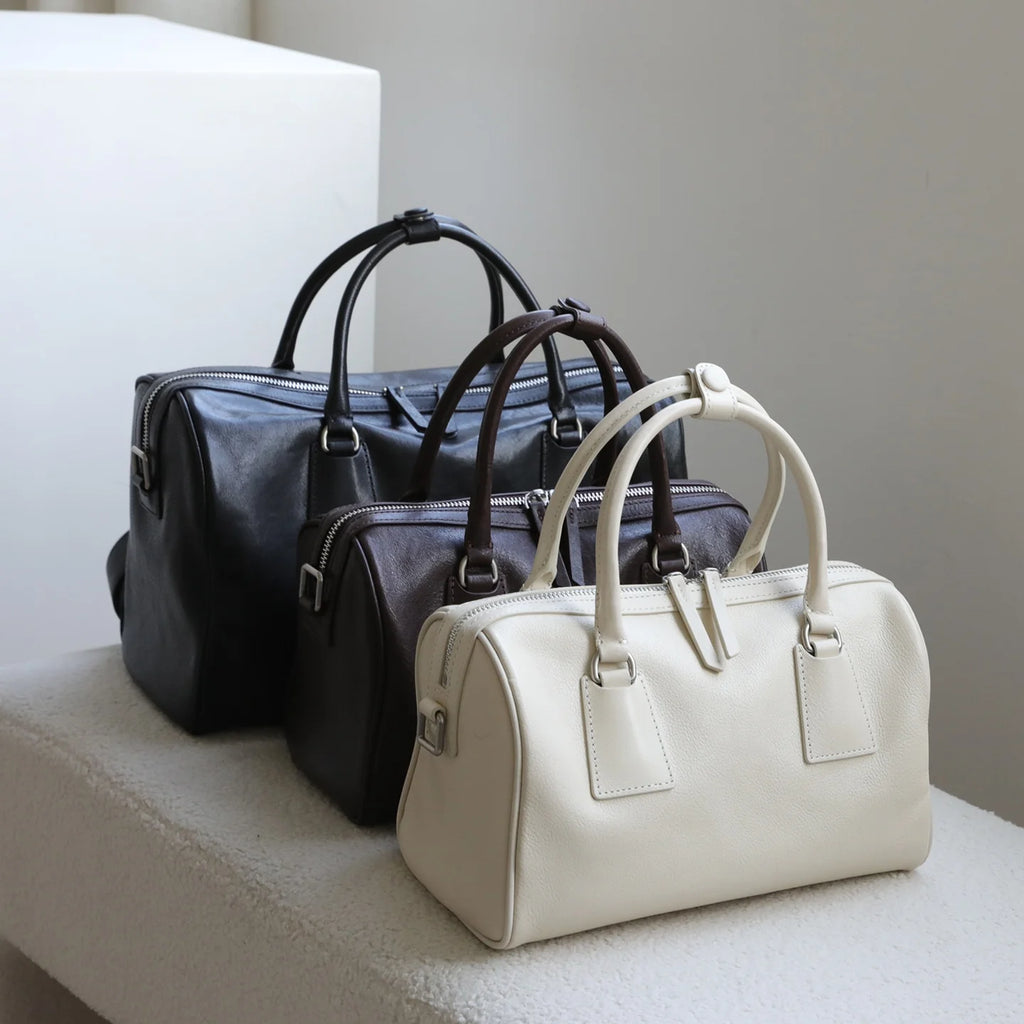 Three minimalist vegetable-tanned leather Boston bags in black, dark brown, and cream with dual zip closures.