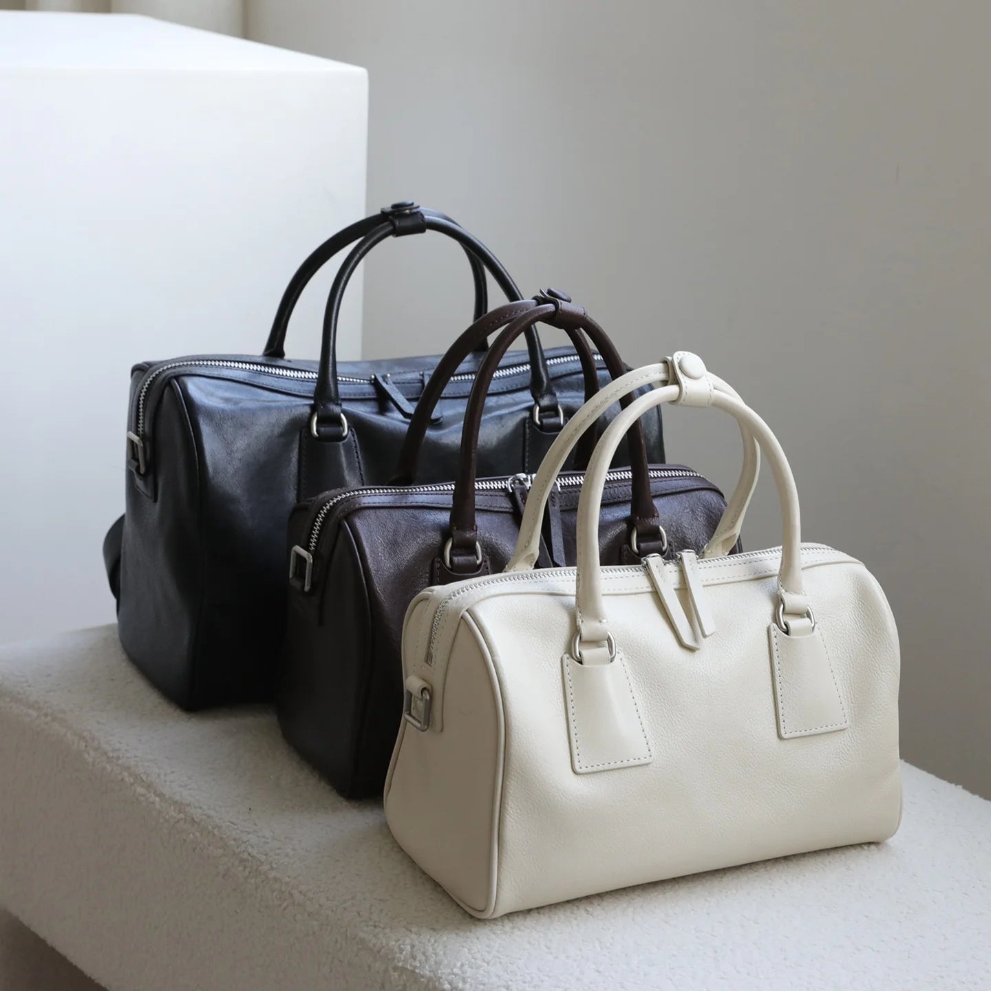 Three minimalist vegetable-tanned leather Boston bags in black, dark brown, and cream with dual zip closures.
