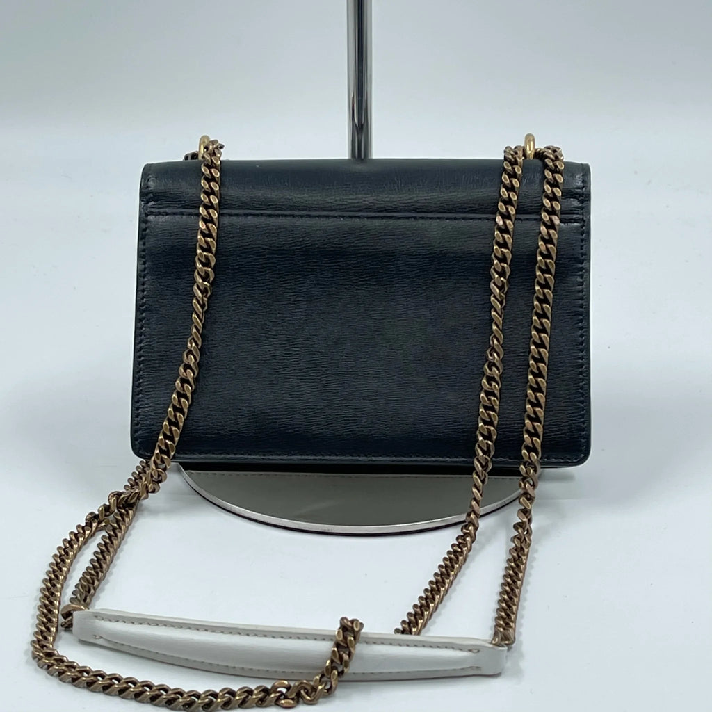 Black leather designer shoulder bag with a snap closure, featuring a two-tone chain strap and white accent piece.