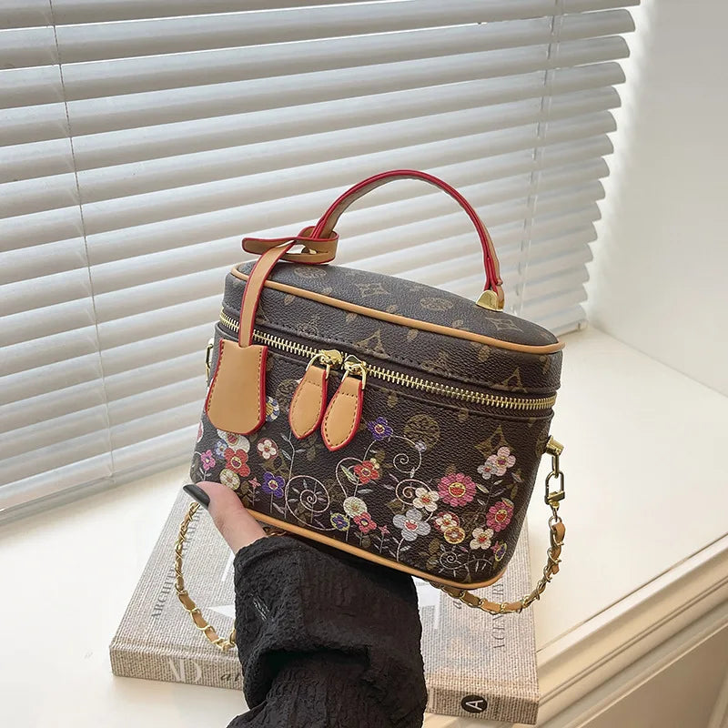 Brown cowhide vanity trunk bag with colorful floral monogram print, gold hardware, and a red-trimmed top handle.
