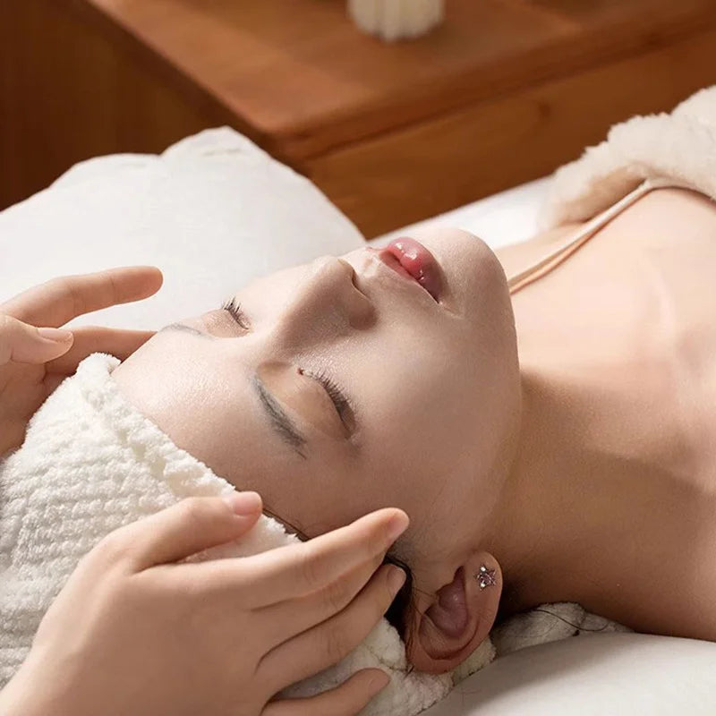 A person with a white headband lying down while receiving a professional facial treatment in a spa setting.
