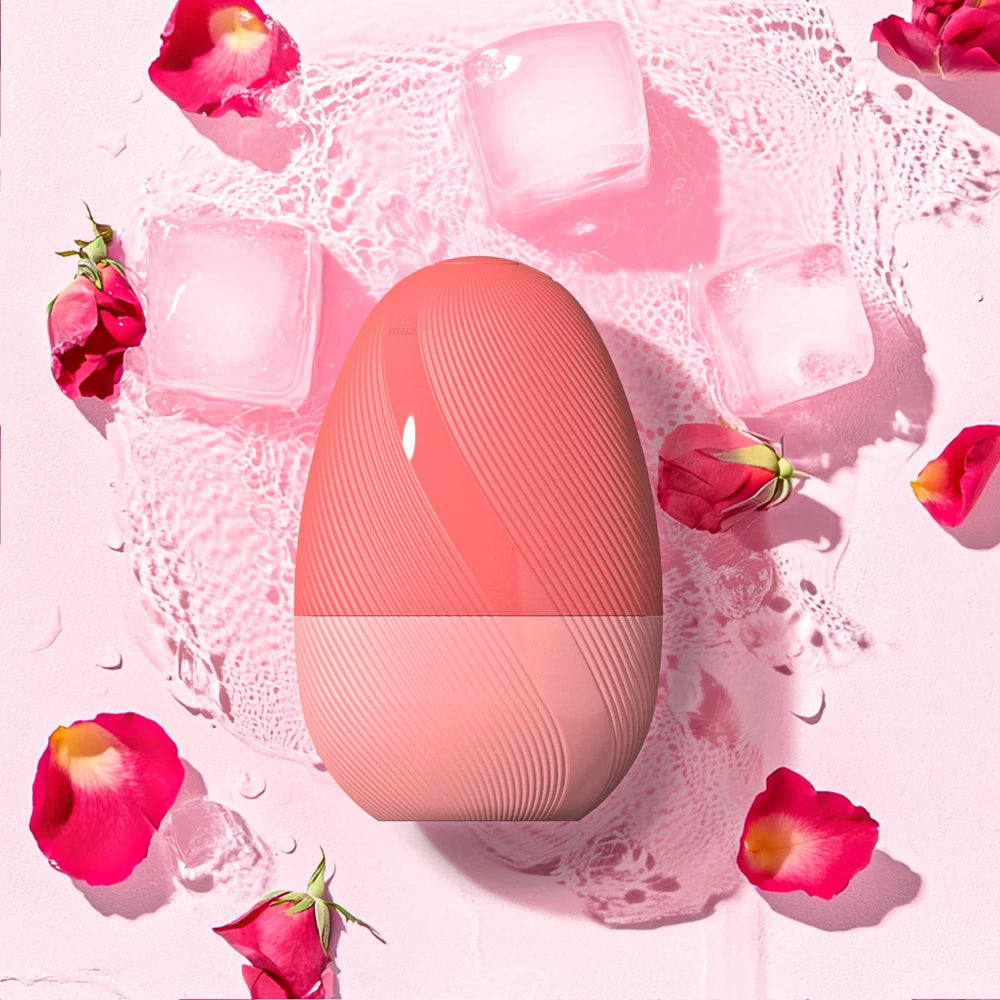 Oval-shaped silicone facial cleansing brush in coral pink, surrounded by ice cubes and rose petals.