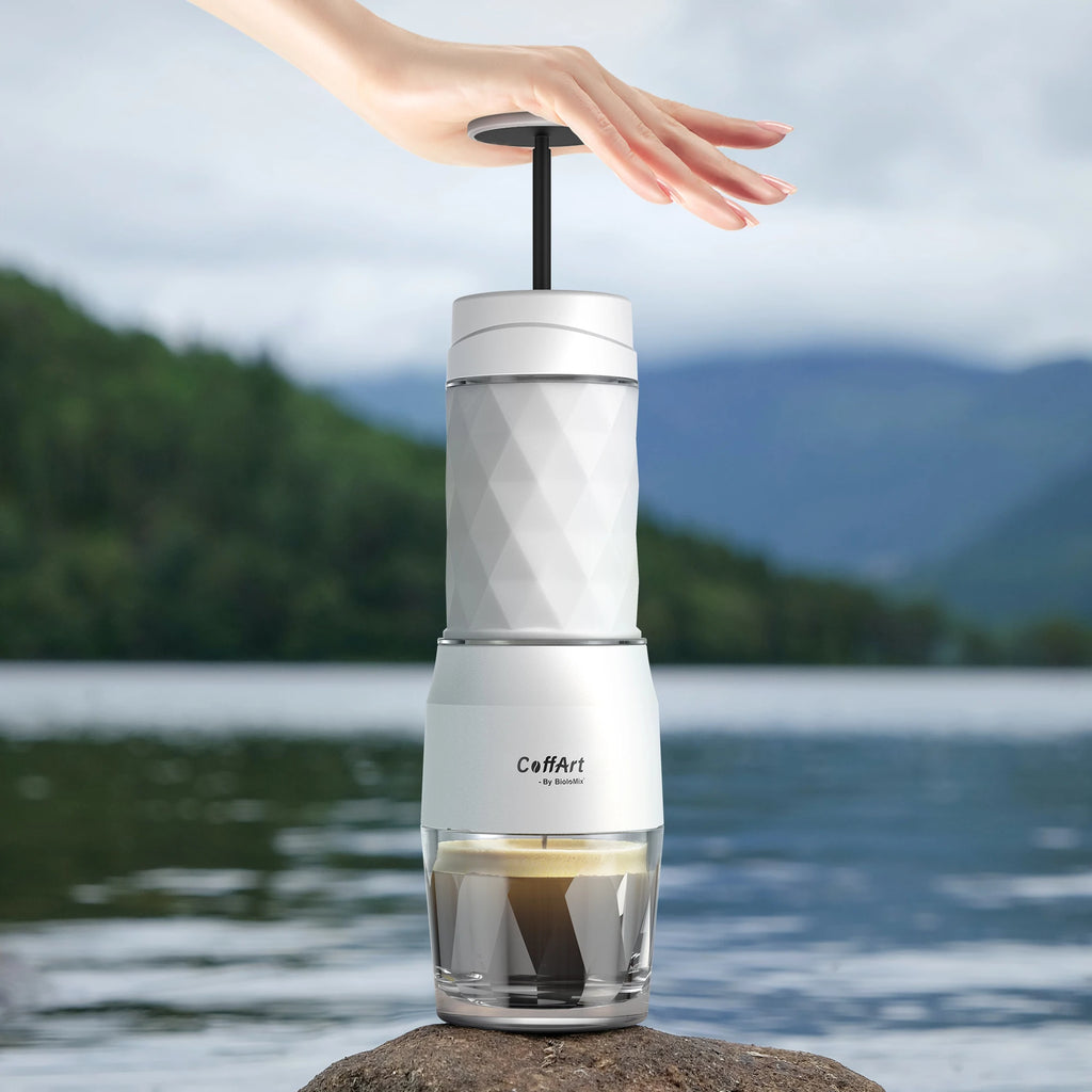 CoffArt by BioloMix portable white manual espresso maker with diamond-textured body brewing coffee on a lakeside rock.