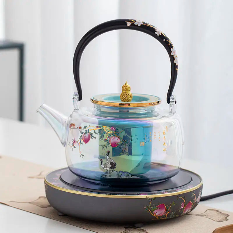 Elegant glass teapot with floral crane print, gold finial, and matching ornate electric warmer base for tea brewing.