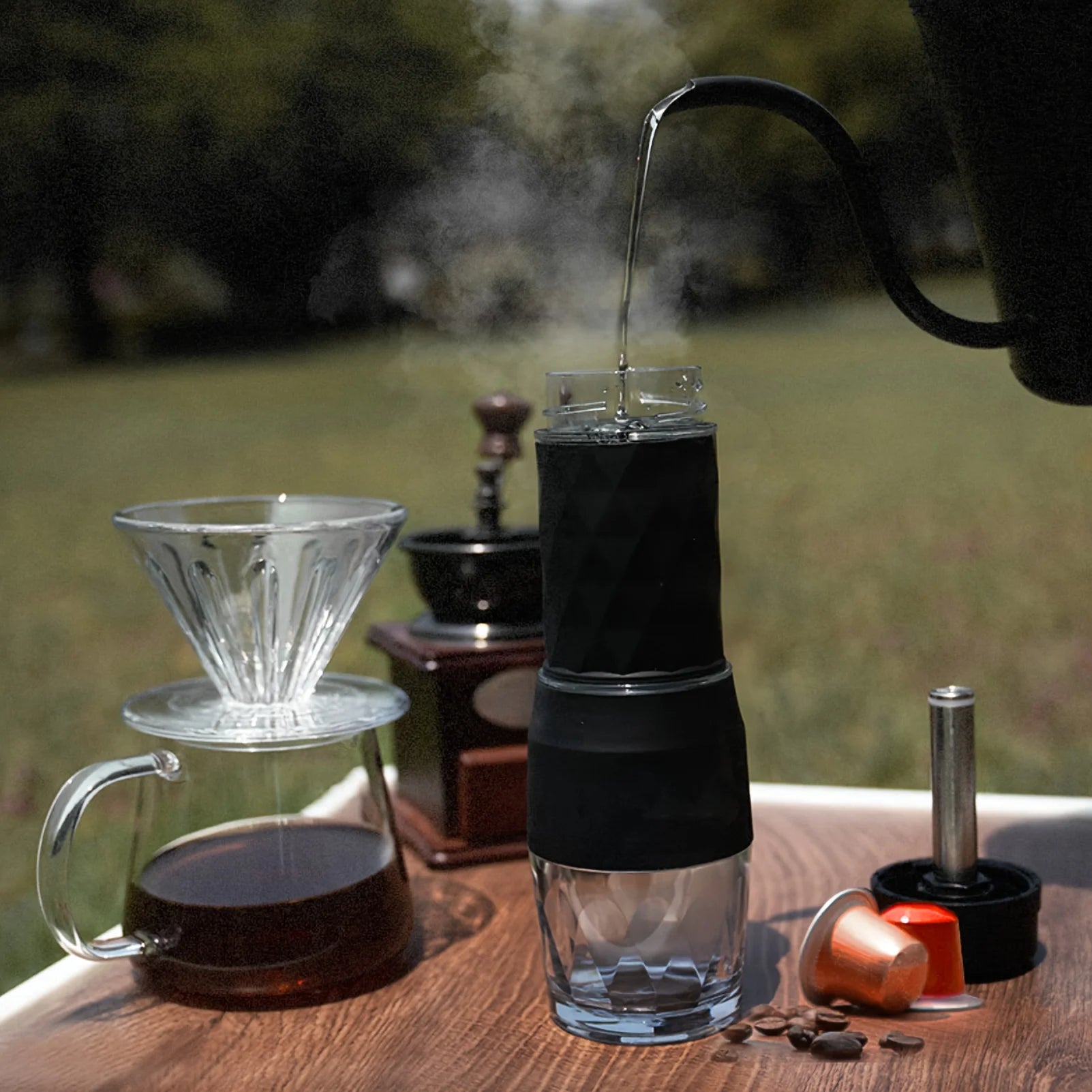 Portable black and glass coffee maker for capsules, pouring hot water with coffee dripper and grinder on table.