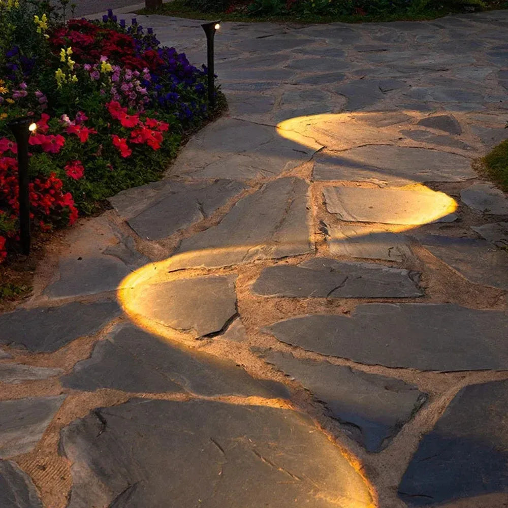 Black solar landscape lights illuminate a stone garden walkway, casting warm, curved light paths.