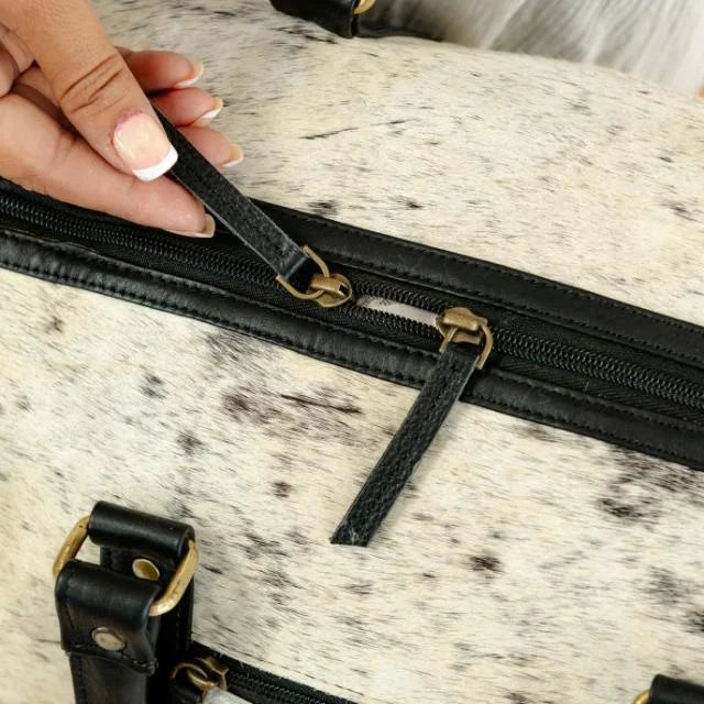 White and black cow hair-on-hide weekender duffel trunk with black leather trim and bronze zipper hardware.