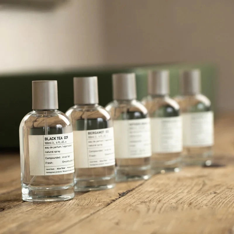 Clear glass spray bottles of perfume with matte grey caps and white rectangular labels, arranged on a wooden surface.