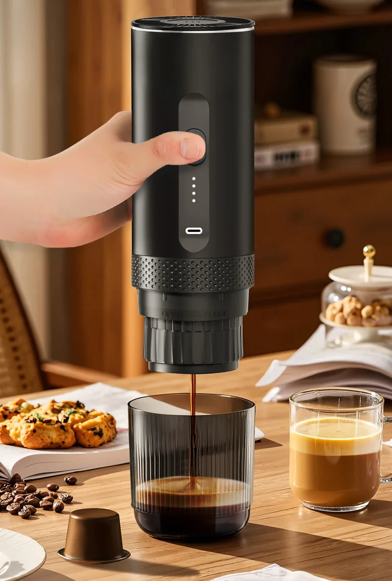 Handheld portable black electric coffee maker pouring espresso into a ribbed glass cup on a wooden table.