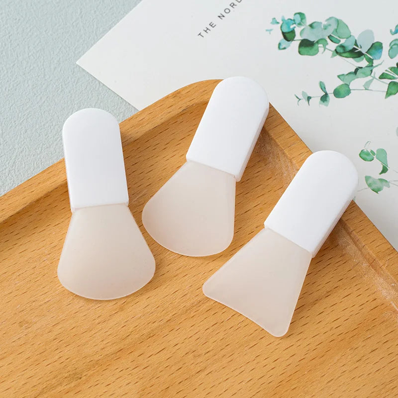 Three silicone facial mask applicators with white handles on a wooden tray.