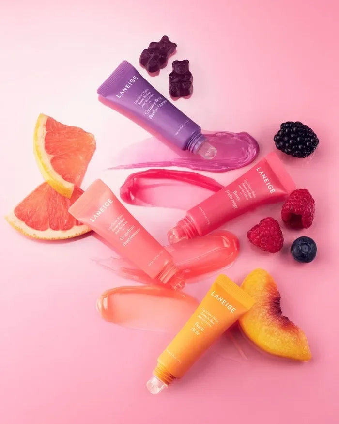 Laneige Lip Glowy Balm set in colorful tubes with glossy swatches, fresh fruit, and candy accents on pink background.