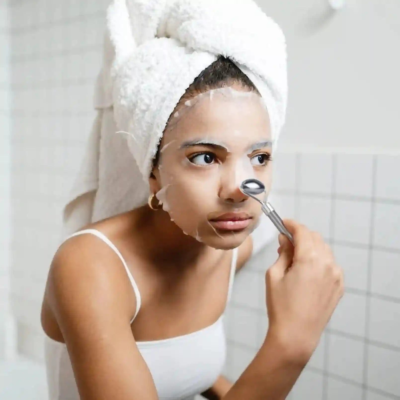 Woman with a towel on her head and a face mask using a smooth, silver stainless steel facial roller on her nose.