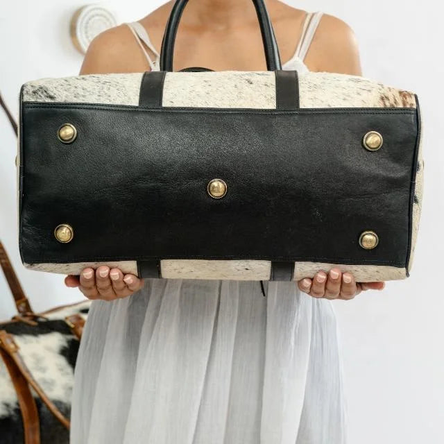 Unisex overnight weekender duffel in black genuine leather and white cow hair-on-hide with brass stud feet.