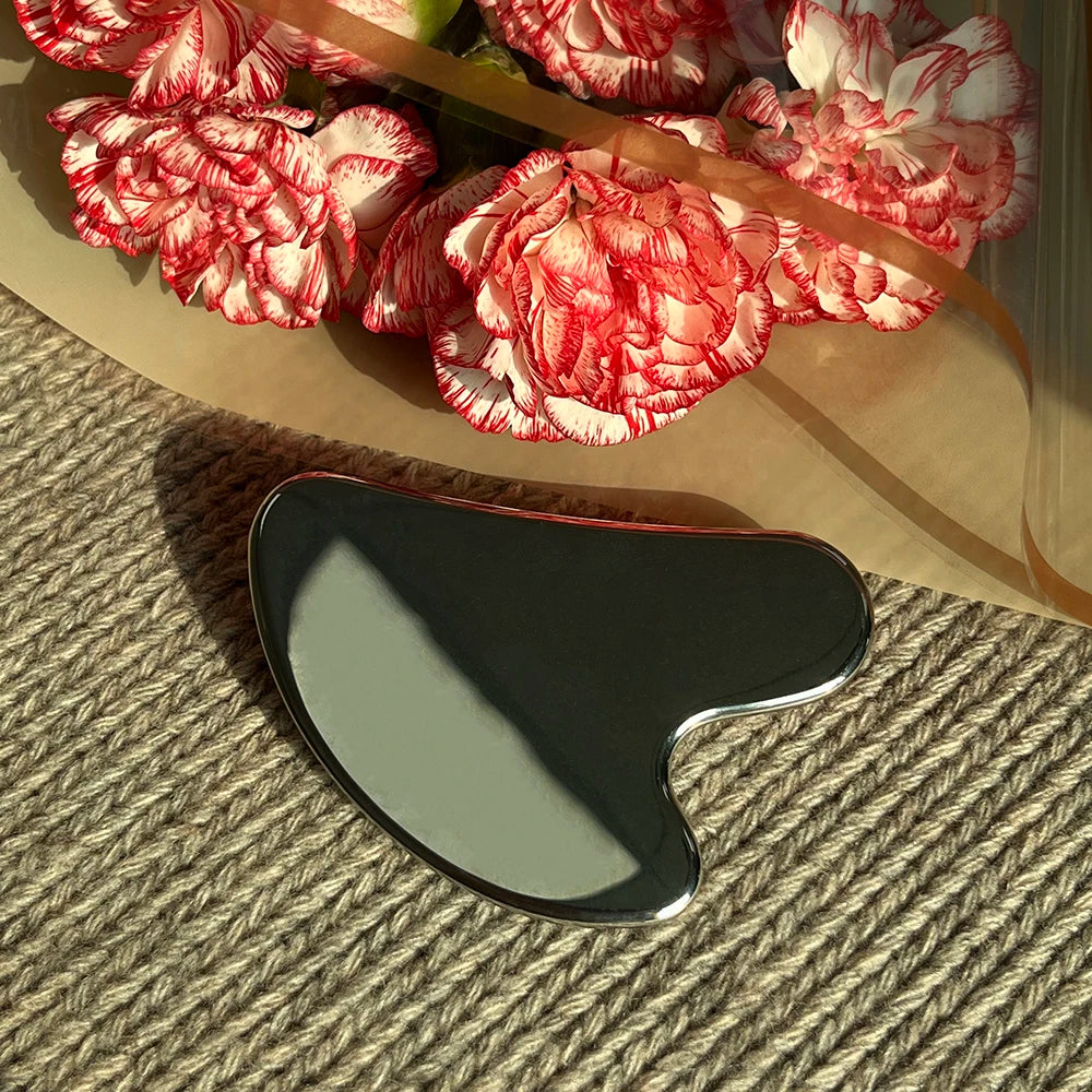 Polished, heart-shaped stainless steel Gua Sha facial tool resting on a textured surface near a bouquet of red flowers.