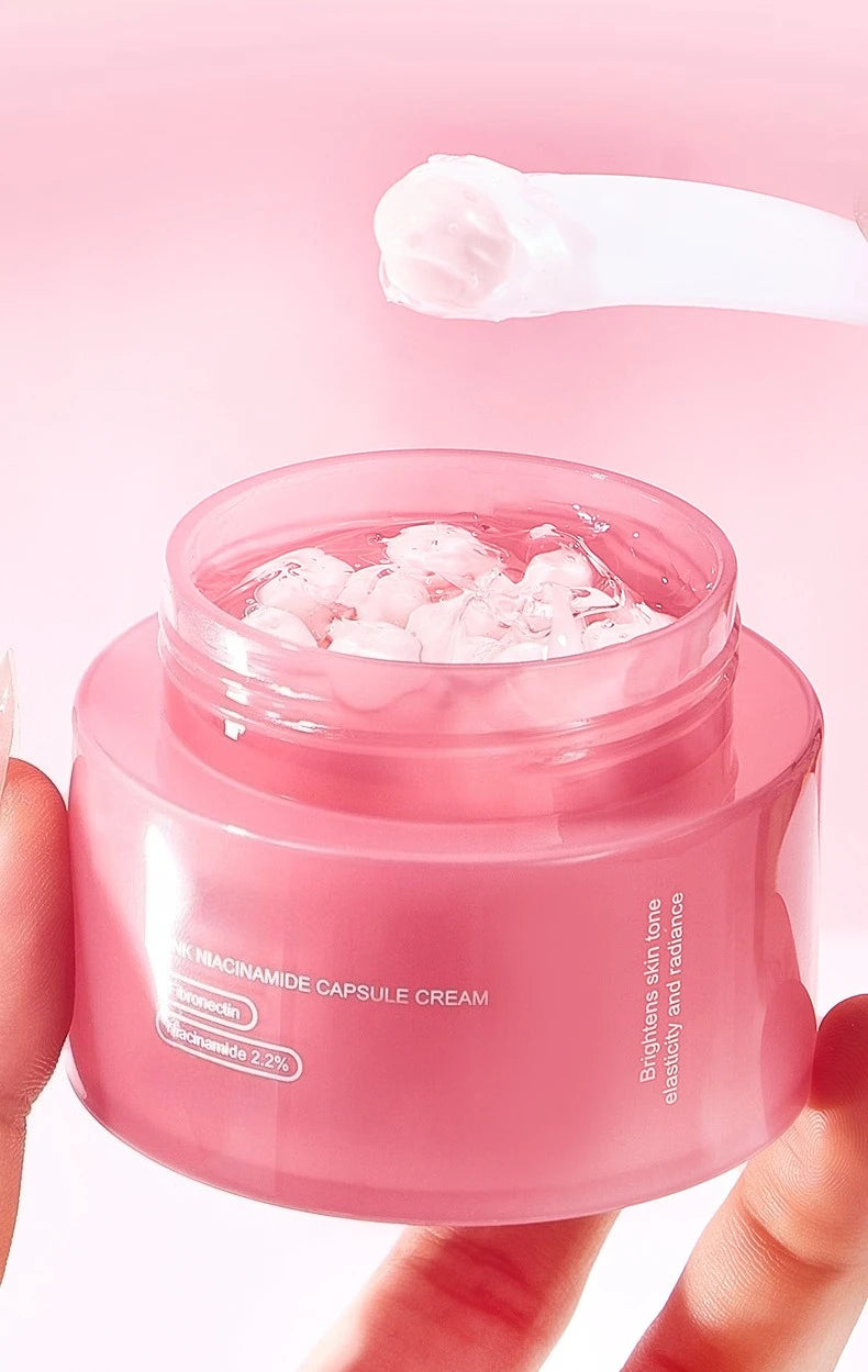 A jar of pink capsule face cream held by fingers, with a small scoop of cream suspended above.