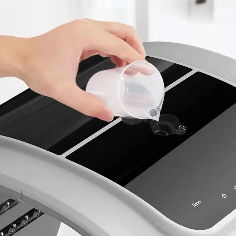 A hand pours water from a small measuring cup into the reservoir of a grey and black LED facial therapy machine.