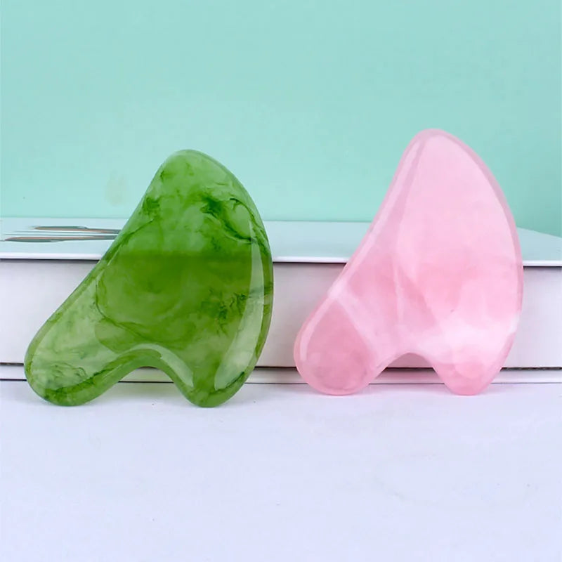 Two smooth, wing-shaped resin gua sha facial massage tools in green and light pink on a white surface.