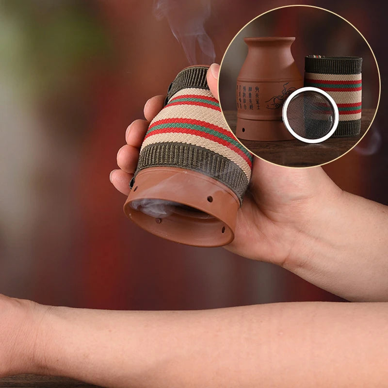 A hand holds a brown ceramic moxibustion cup wrapped in striped fabric, releasing wisps of smoke onto an arm.