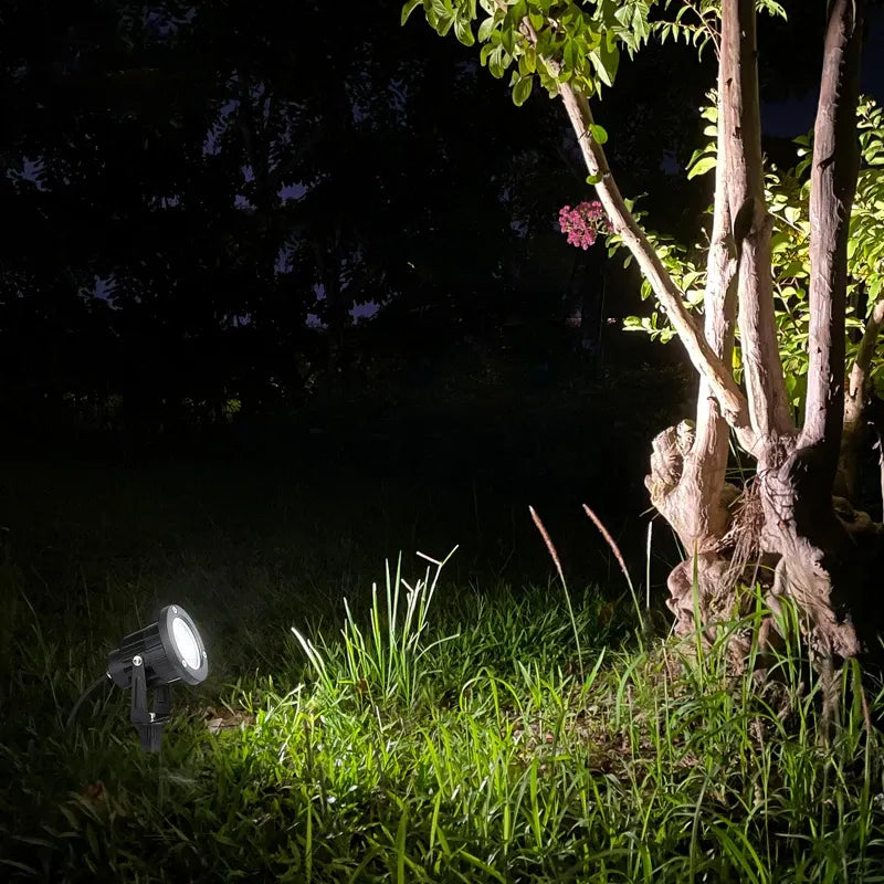 Two outdoor LED spike lights on grass beside tree, casting warm white light