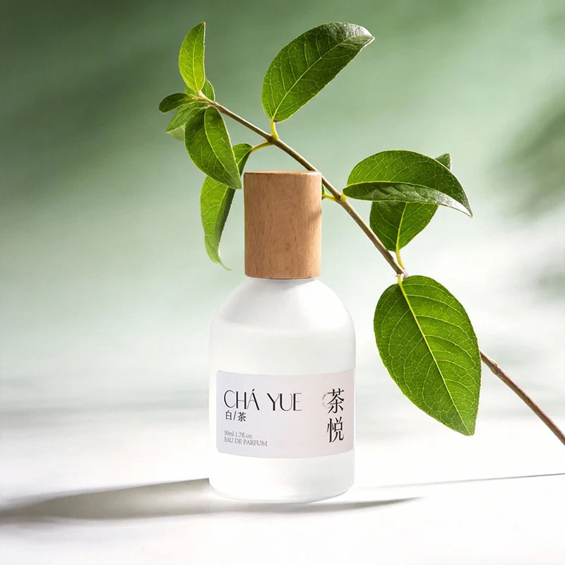 Frosted glass white tea perfume bottle with wooden cap, minimalist label, and green leaves on a light green background.