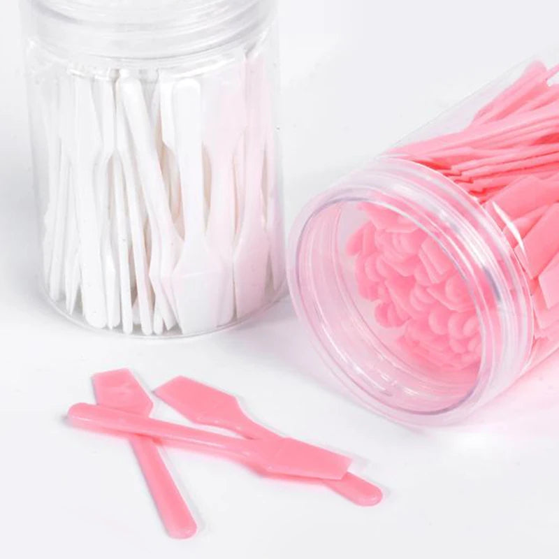 Two clear jars filled with small white and pink plastic cosmetic spatulas, with a few loose pink ones in front.