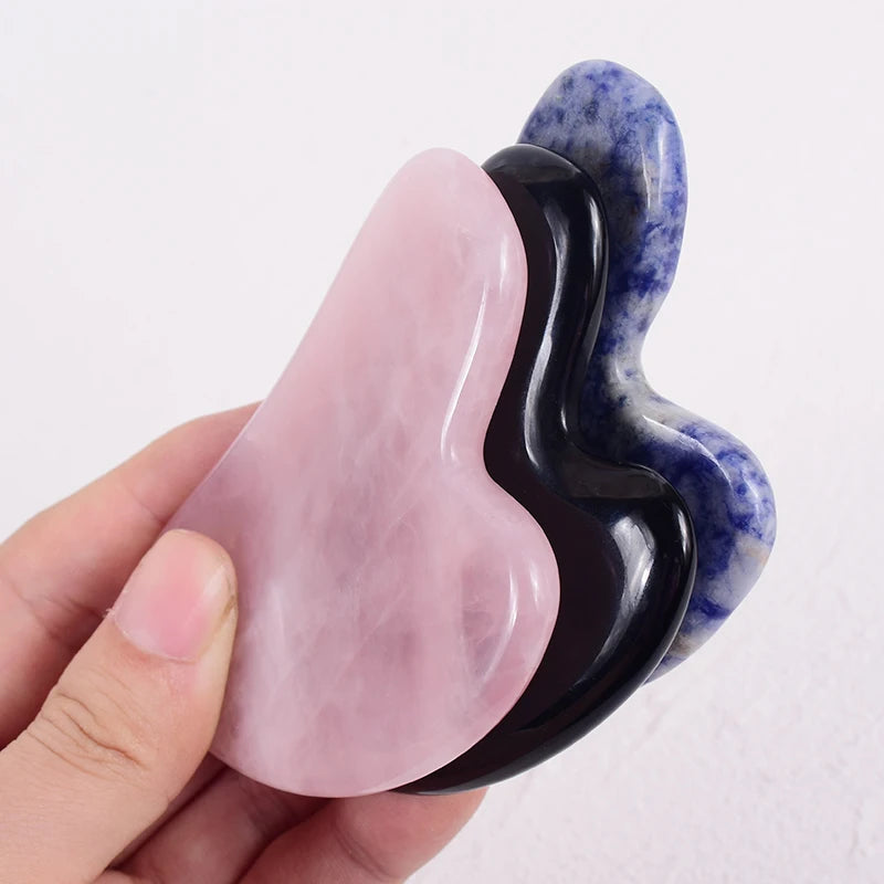 A hand holding three smooth, heart-shaped gua sha stones: one pink rose quartz, one black obsidian, and one blue stone.