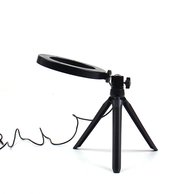 Small black ring light on a foldable plastic tripod stand with a power cord, used for tabletop video lighting.