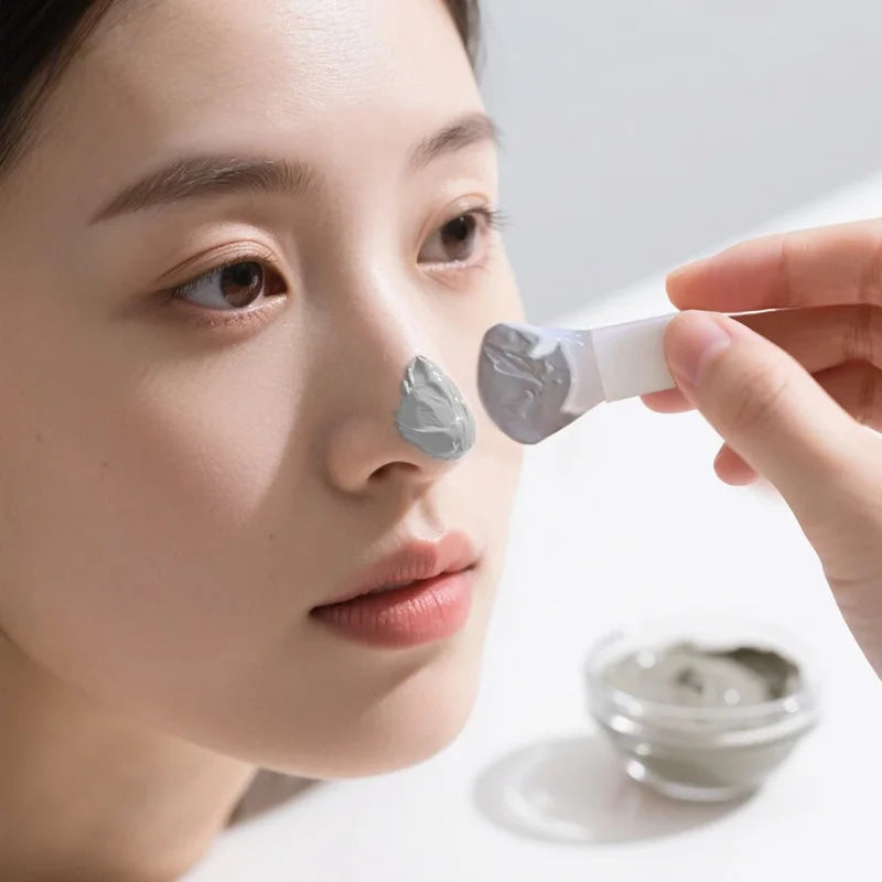 A person uses a white silicone applicator to spread gray mud mask onto the tip of their nose.