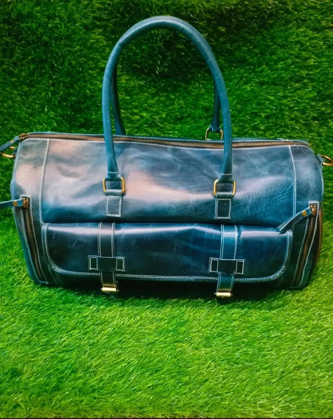Blue full-grain leather duffle bag with side pockets, brass hardware, and top handles. Luxury designer travel bag.