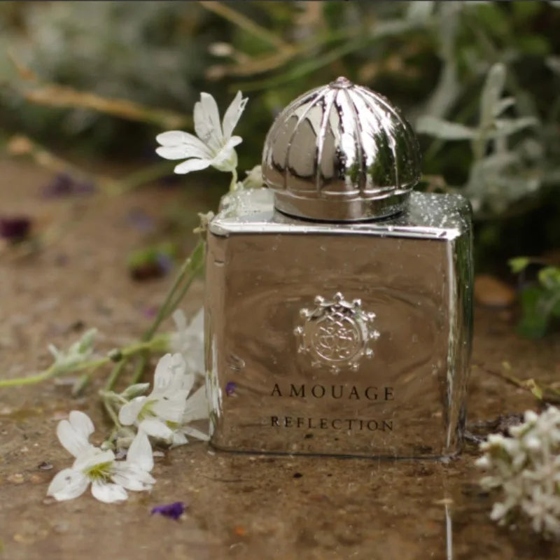 Metallic silver Amouage Reflection perfume bottle with a dome cap, surrounded by white flowers on a wet surface.