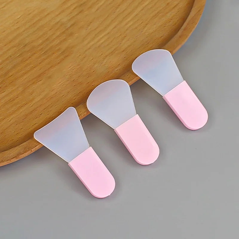 Three silicone facial mask applicators with opaque white flexible heads and solid pink handles, angled on a tray.