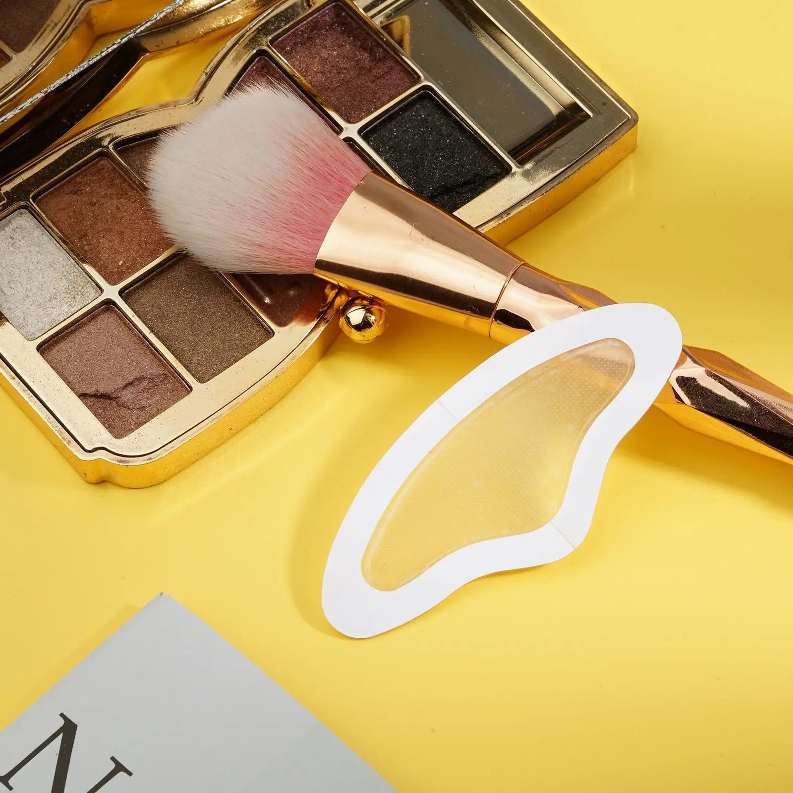 A translucent, boomerang-shaped microneedle forehead patch rests on a yellow surface next to a makeup brush and palette.