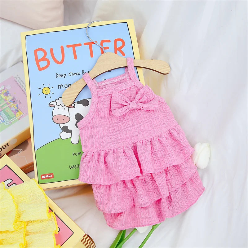 Pink sleeveless dog dress in crinkled fabric with a front bow and three-tiered ruffled skirt on a wooden hanger.